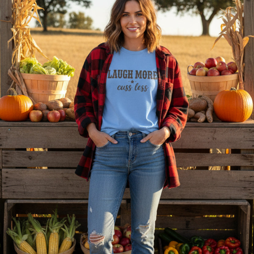 A light blue short-sleeve t-shirt with the text 'LAUGH MORE cuss less' on a woman by a farm stand