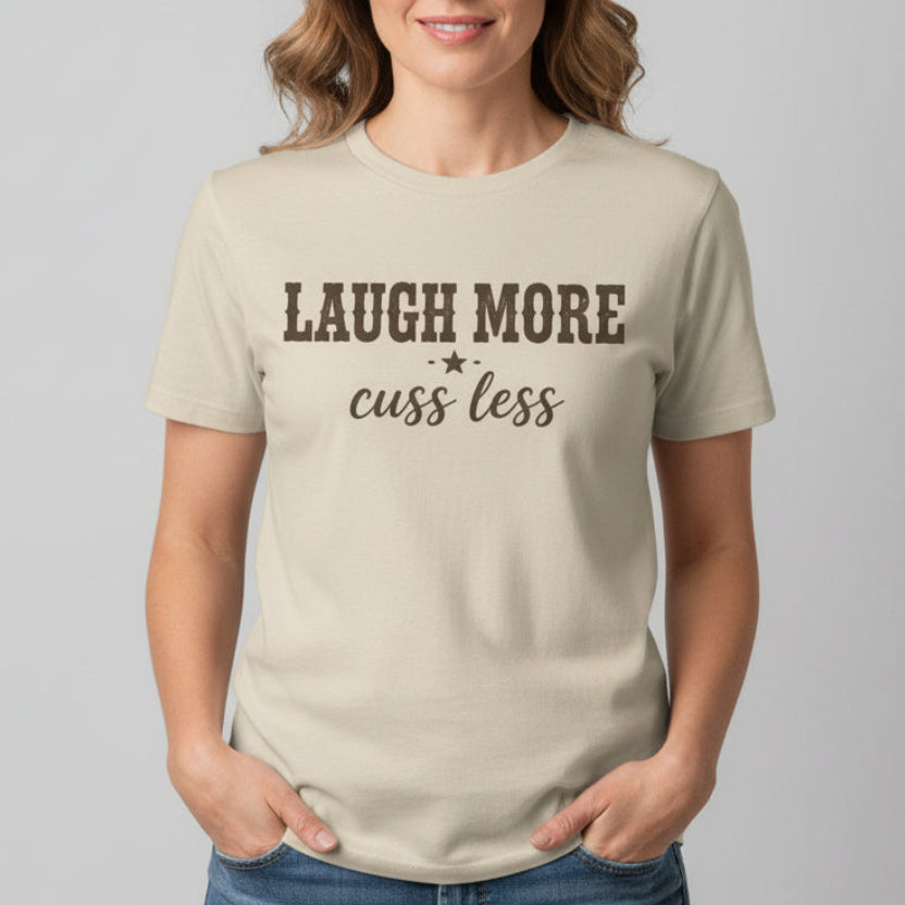 A beige crewneck t-shirt with the text 'LAUGH MORE cuss less", displayed on a clean white background.