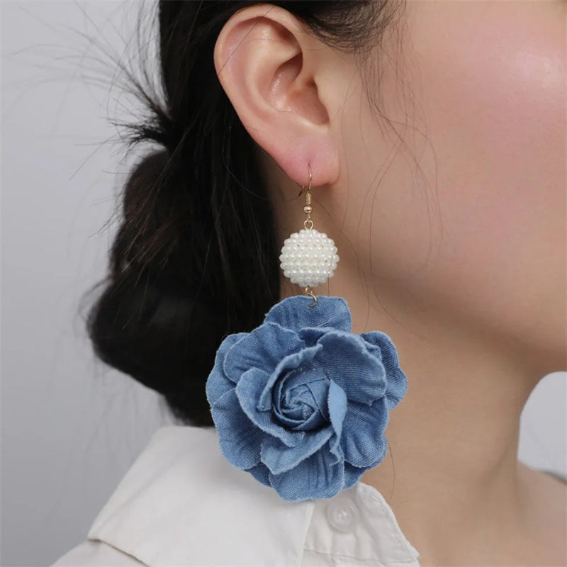 Blue flower earring with pearl-like detail worn by a person against a neutral background