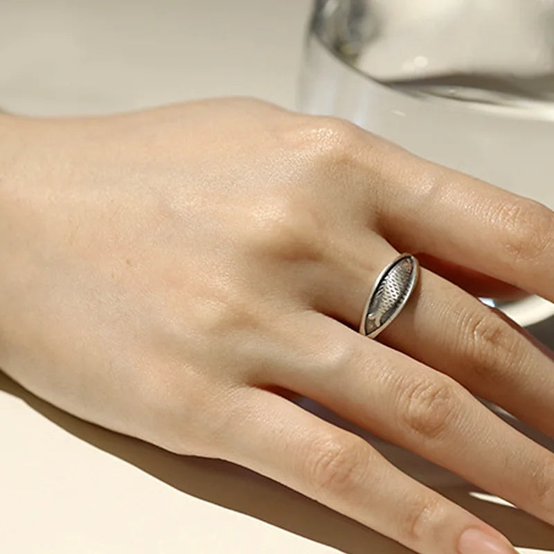 Hand wearing a silver ring with a textured fish design on a neutral background