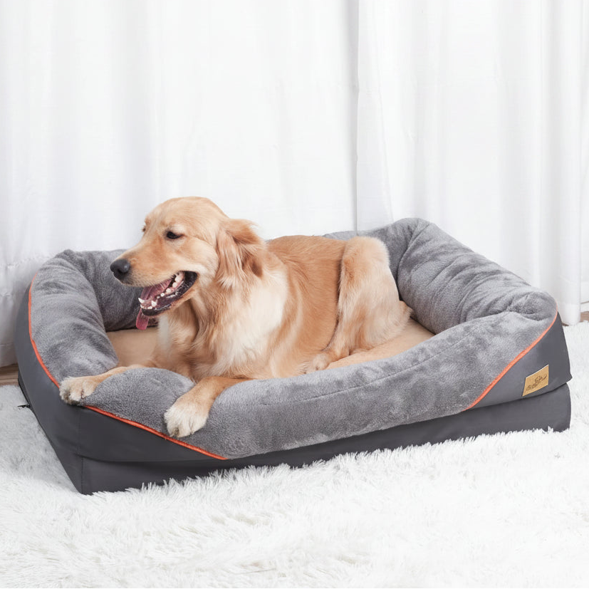 A golden retriever dog rests inside a plush gray pet bed with orange piping and a small gold brand tag, set on a white shag rug in front of white curtains.