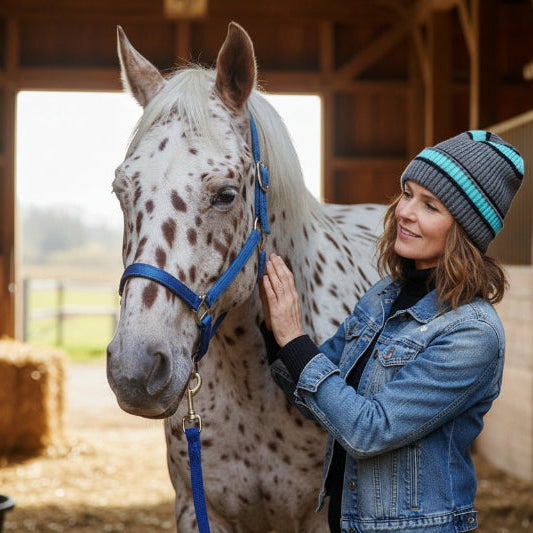 Woman in a blue denim jacket and gray knit hat standing next to a white horse with brown spots in a stable.