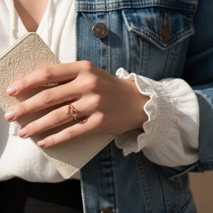 Hand with rose gold heart knot ring holding a beige embossed clutch with a denim jacket and white blouse in the background