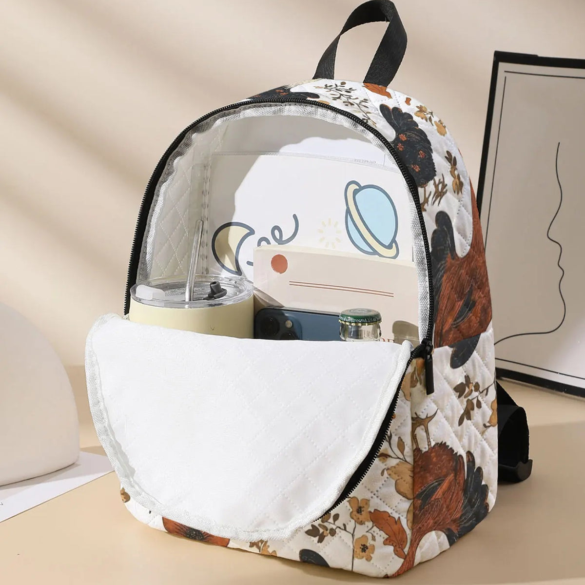 A patterned backpack with a clear front window and white front pocket, shown on a beige tabletop with a cup inside and framed wall art in the background.