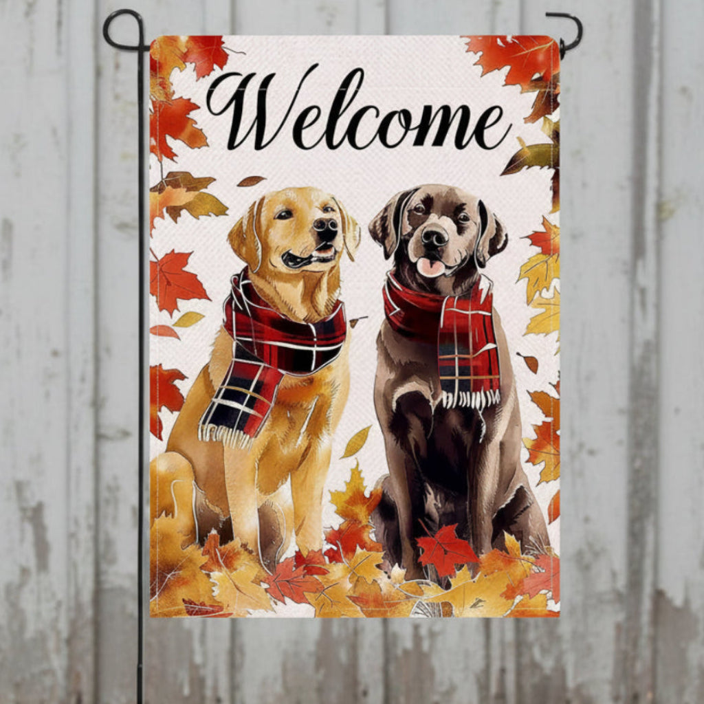 A decorative garden flag with a 'Welcome' message in black script, surrounded by autumn leaves and featuring two dogs wearing red plaid scarves. The flag is displayed on a black metal stand against a white background.