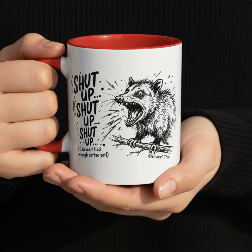 A white ceramic mug with a bright red interior, featuring a black line art illustration of a snarling opossum and the text 'SHUT UP.