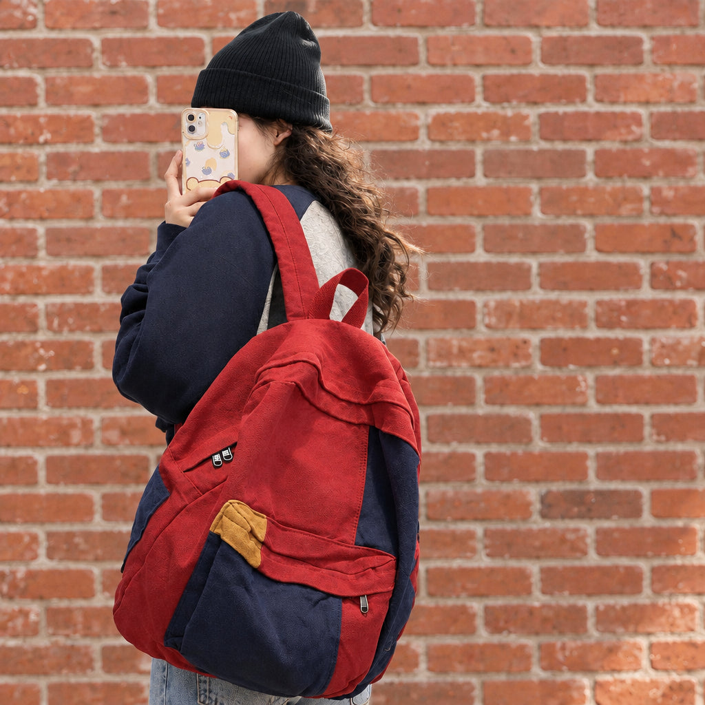 A person standing outdoors in front of a high school building, wearing a red and navy backpack with a small yellow pocket detail. The person is seen from the back, holding a phone.