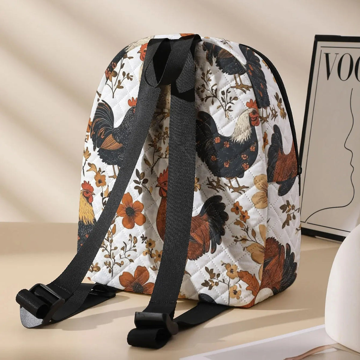 A quilted canvas backpack with a black and brown rooster and floral pattern on a white background, featuring black adjustable shoulder straps, styled on a beige surface next to a framed print.