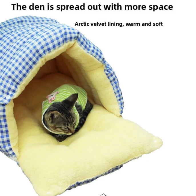 A cat wearing a green outfit lies inside a soft cave-like pet bed with a blue-and-white gingham exterior and pale yellow interior. Text above reads, 'The den is spread out with more space' and 'Arctic velvet lining, warm and soft.