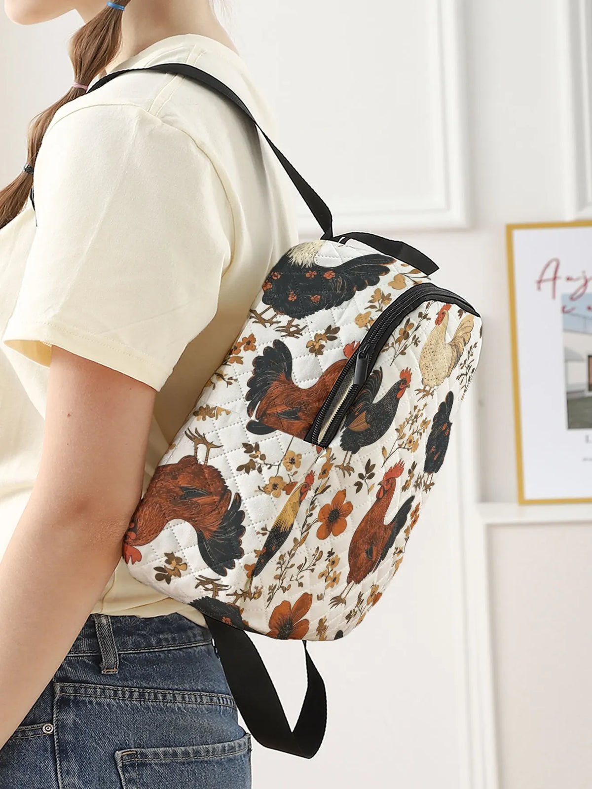 A person wearing a cream t-shirt and blue jeans carries a small backpack with a vintage-style rooster and floral print on a white background. The backpack features black straps and trim, shown in a bright indoor setting.